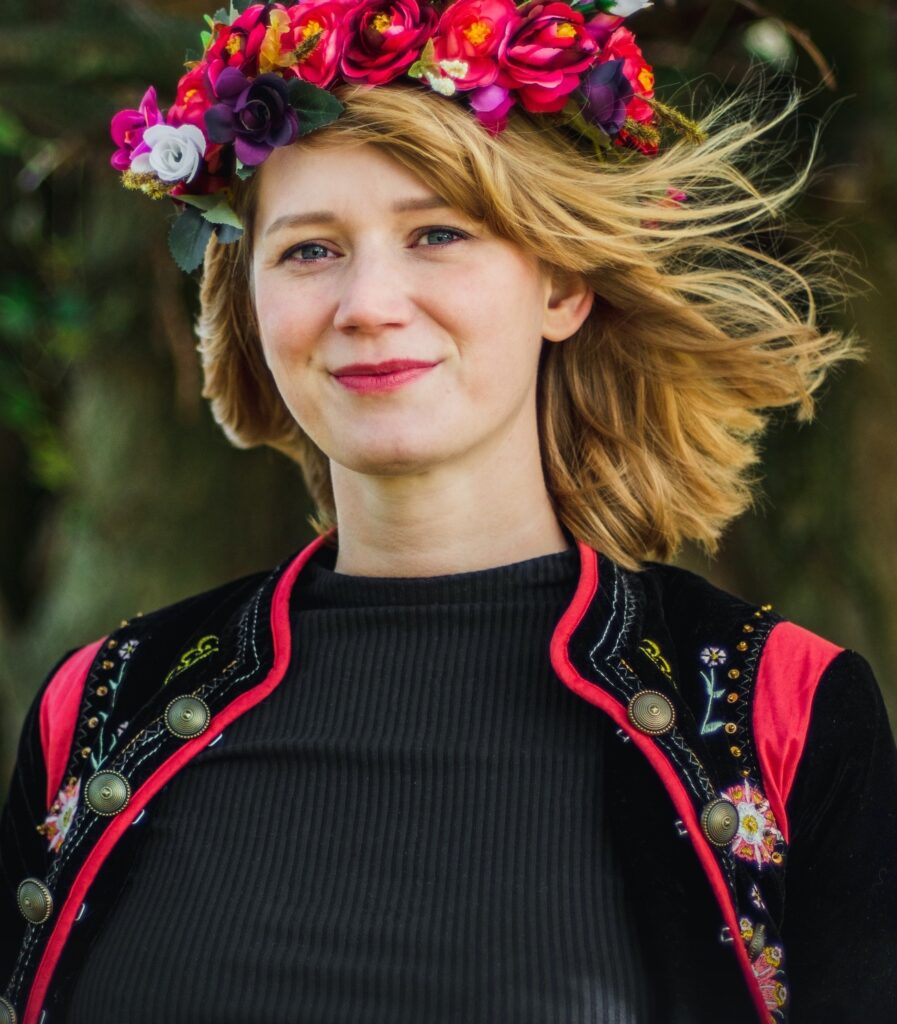photo of a blonde woman in a black t-shirt, black and red cardigan, wearing a flowercrown of red flowers. she is smiling